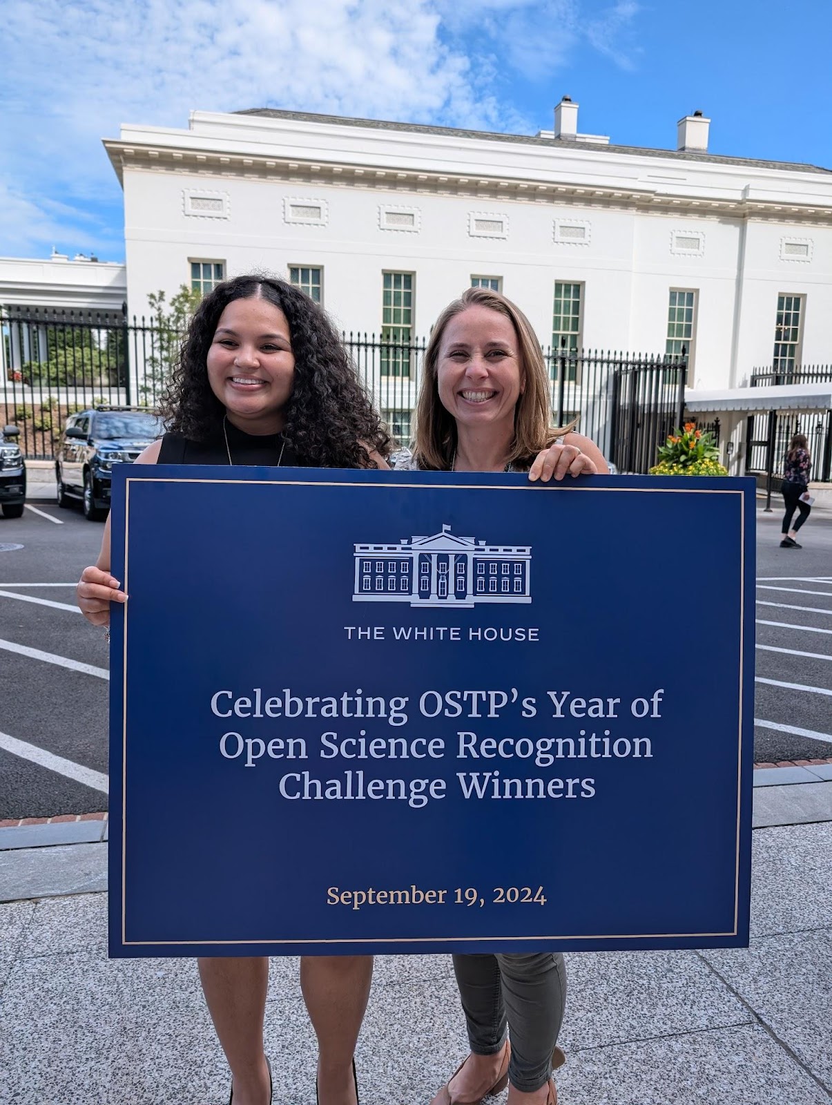 a Black woman and a white woman smiling standing in front of a large single storey building holding a large sign with logo of The White House and text 'Celebrating OSTP's Year of Open Science Recognition Challenge Winners dated September 19, 2024'