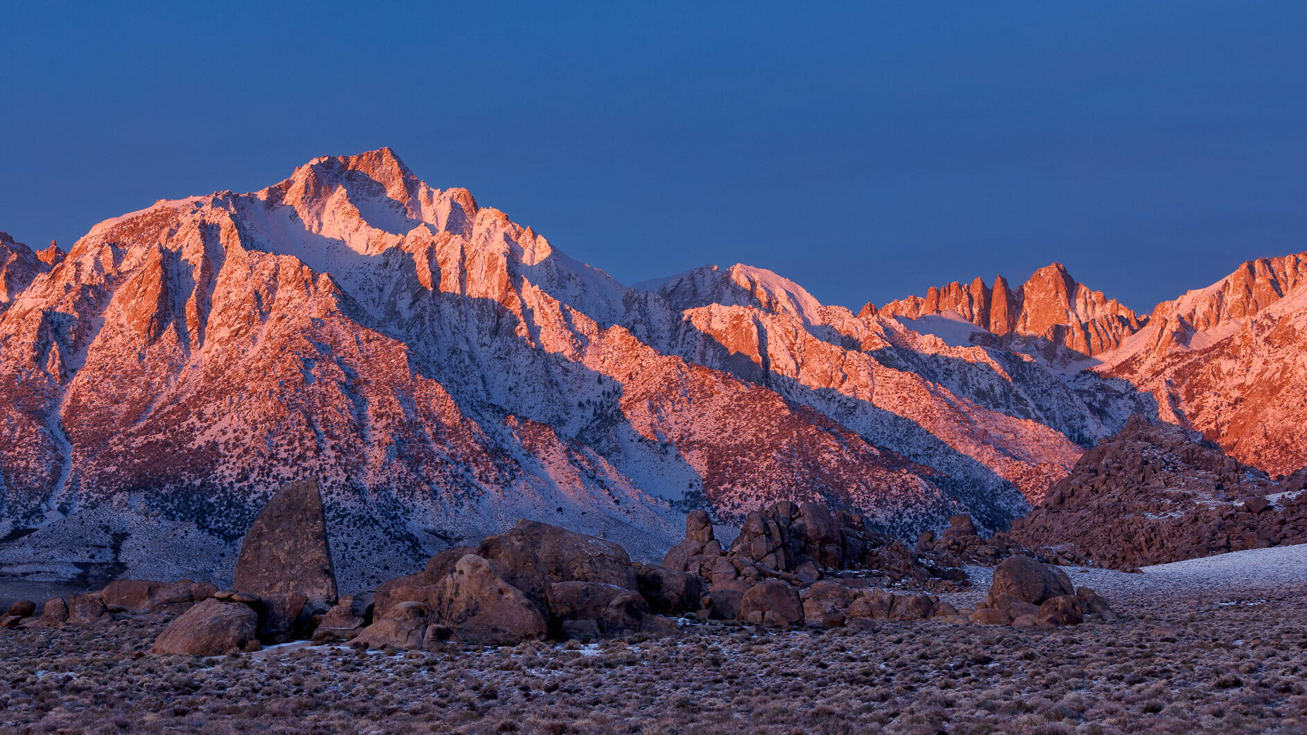 Mount Whitney sunrise. Deep blue clear sky. Reddish mountains with a dusting of snow, with each sharp ridge casting a shadow on the next ridge to the right. Boulders in the foreground.