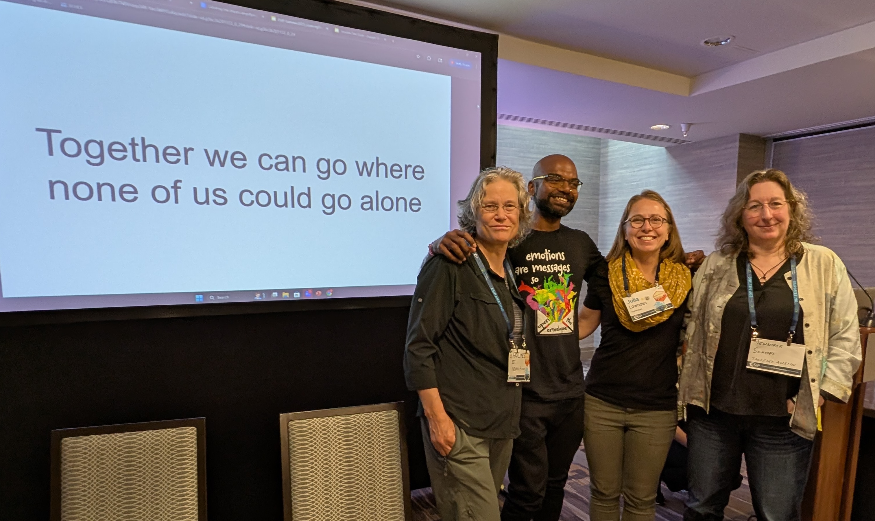 four people smiling standing by a podium with the screen saying 'Together we can go where none of us could go alone'