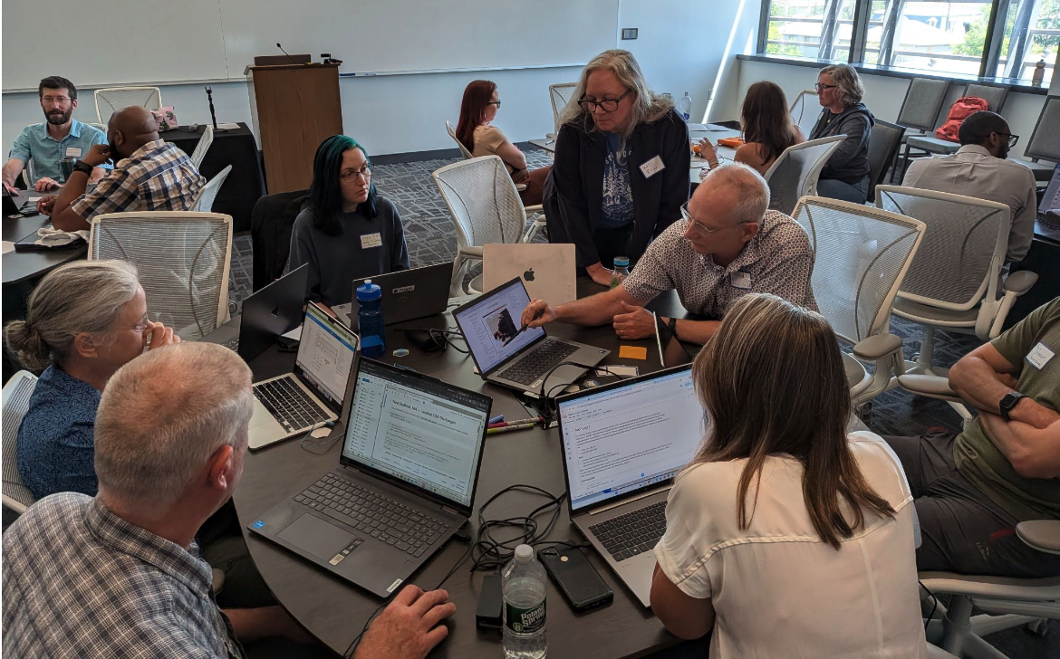 A group of six people sit or stand around a round table with laptop screens open. One person points to their screen as others watch or listen.