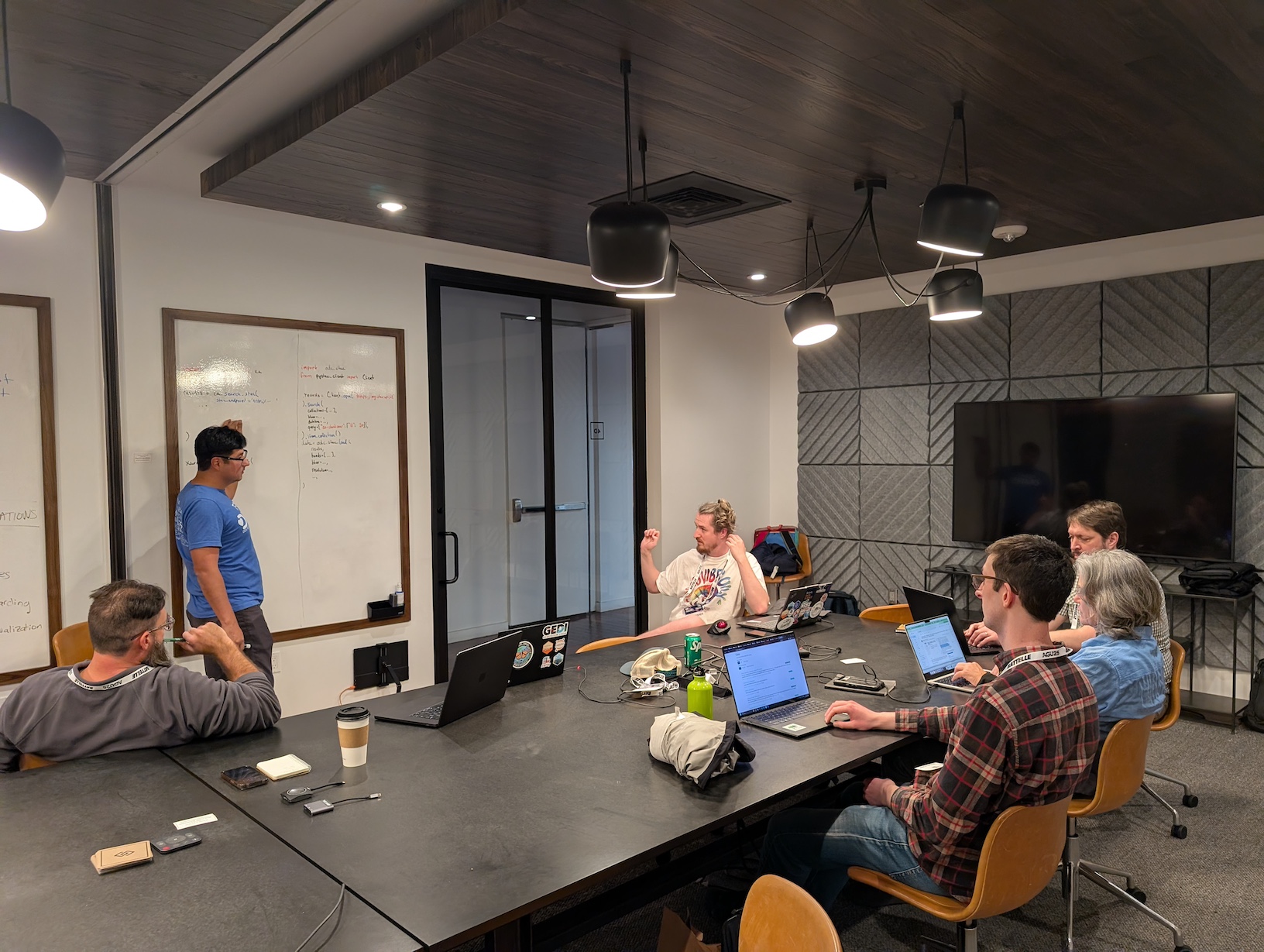 5 people seated around a rectangular table with laptops open looking a a person standing pointing to a whiteboard
