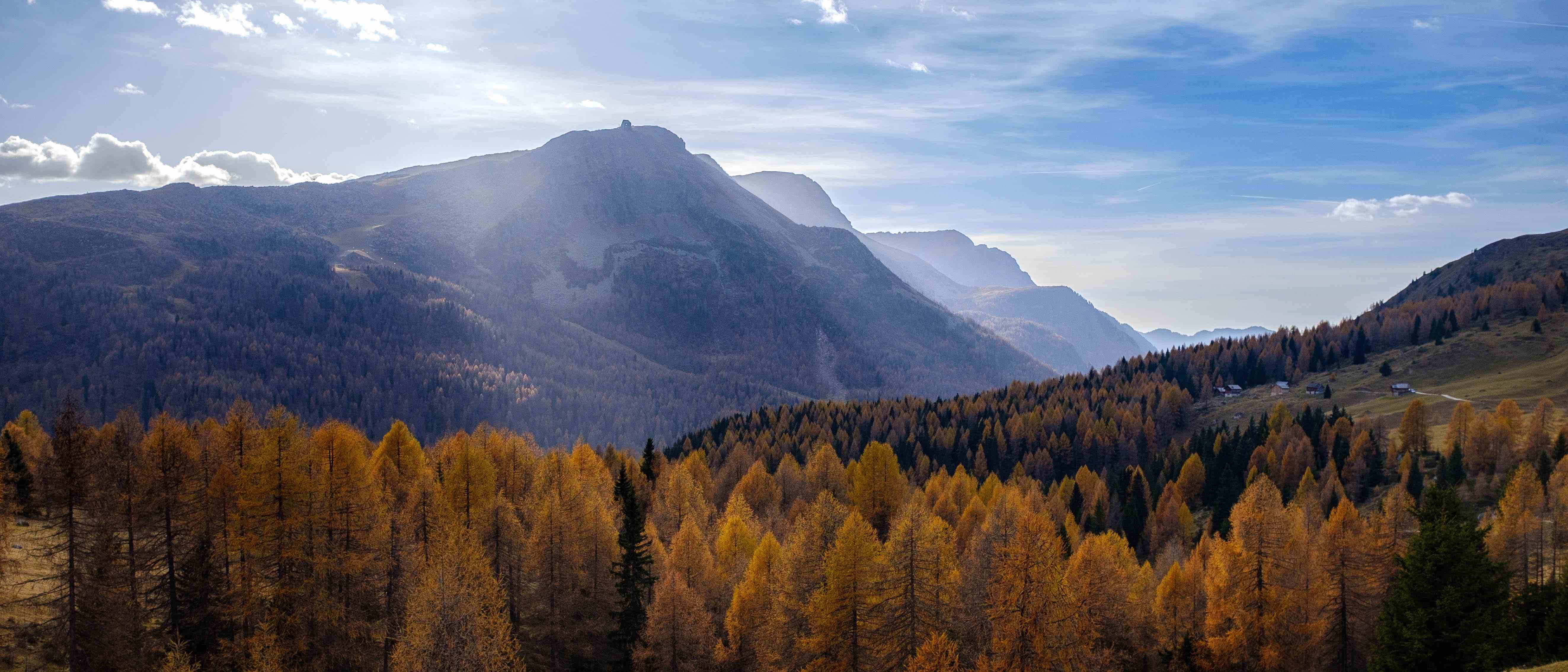 photo of yellow trees in the foreground and tall mountains in the background with light through broken clouds