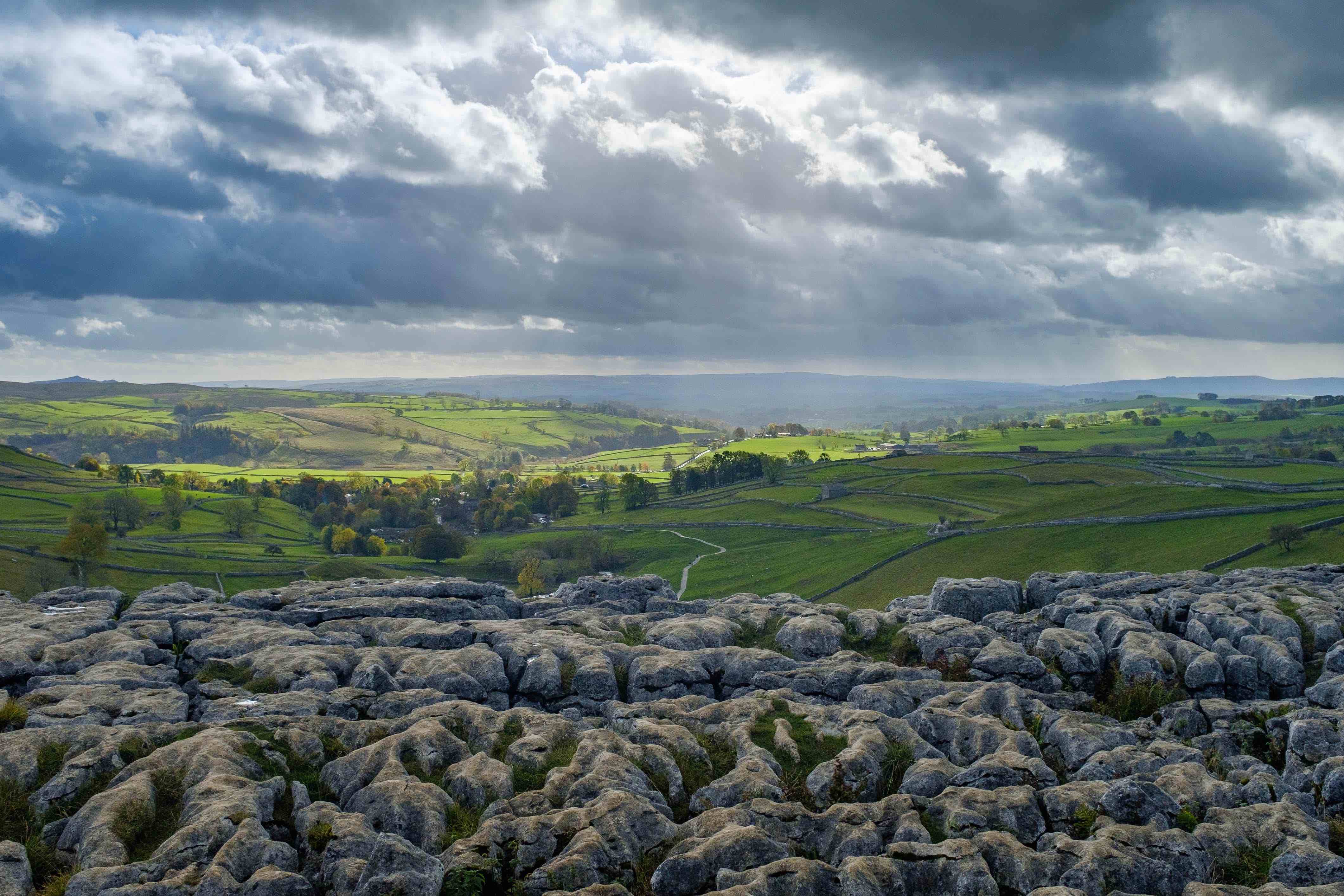 grey rocks that create a limestone pavement geological feature in the foreground with green hills in the background with sun through broken clouds